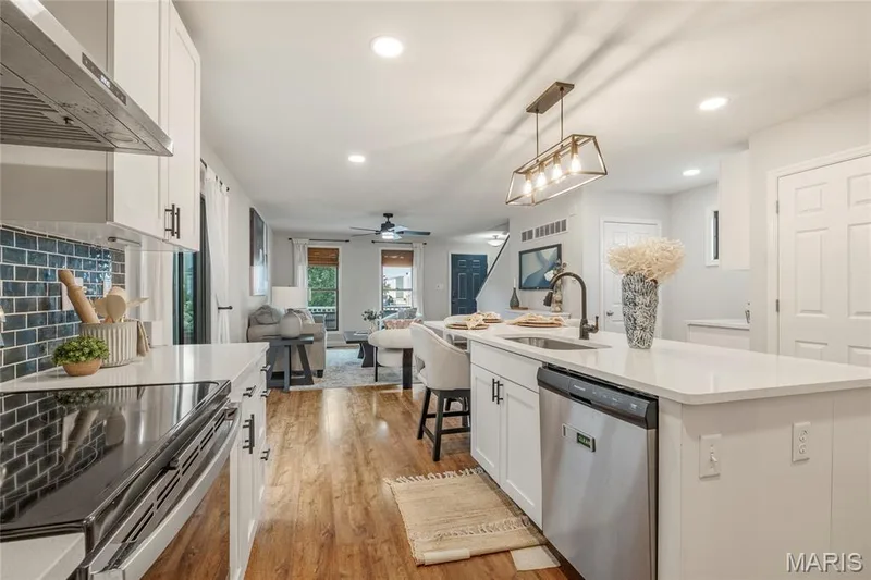 Open-concept kitchen featuring white cabinetry, light countertops, subway tile backsplash, and stainless steel appliances, with wood-finish flooring extending into the living area — 3429 Sublette Street, St Charles, MO 63301