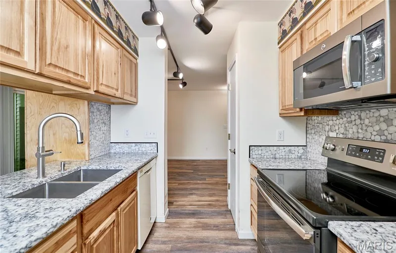 Galley kitchen featuring wood cabinetry, granite-finish countertops, stainless steel appliances, and a mosaic tile backsplash — 146 Old Buggy Court #A, St Charles, MO 63304