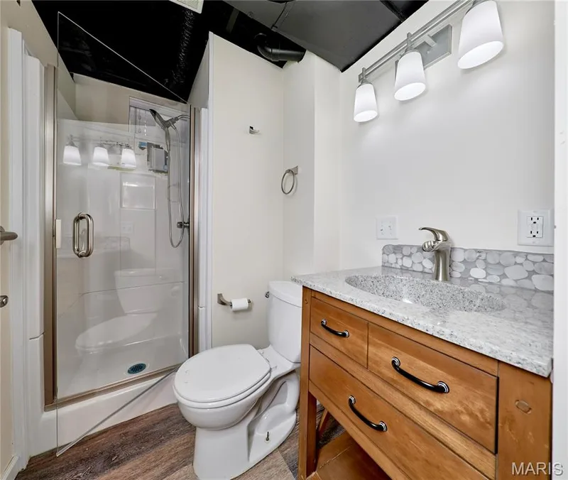 Lower level bathroom with walk-in shower with clear glass enclosure, wood-finish flooring, and a vanity featuring a light-toned countertop, pebble-style backsplash, and wood cabinetry — 146 Old Buggy Court #A, St Charles, MO 63304