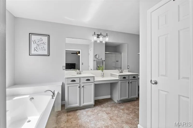 Primary Suite Bathroom featuring a white built-in bathtub, a double vanity with light gray cabinetry, and a walk-in shower with a glass enclosure — 921 Molloy Drive, O'Fallon, MO 63366