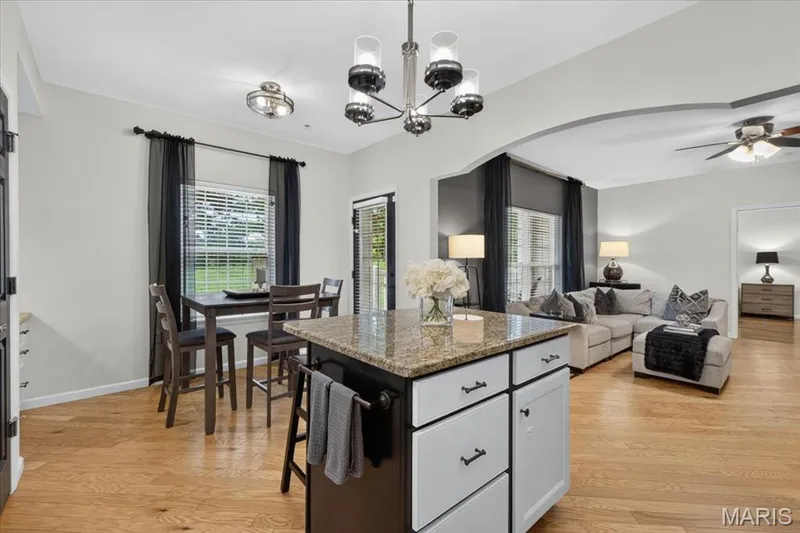 Open-concept living area featuring light wood-finish flooring, a kitchen island with a stone countertop, recessed ceiling details, a modern chandelier, and ample natural light — 5 Monarch Trace Court #108, Chesterfield, MO 63017
