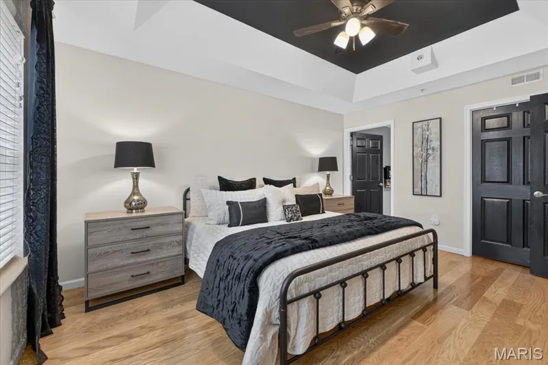 Bedroom featuring light wood-finish flooring, a tray ceiling with a dark accent panel and ceiling fan, and panel doors — 5 Monarch Trace Court #108, Chesterfield, MO 63017