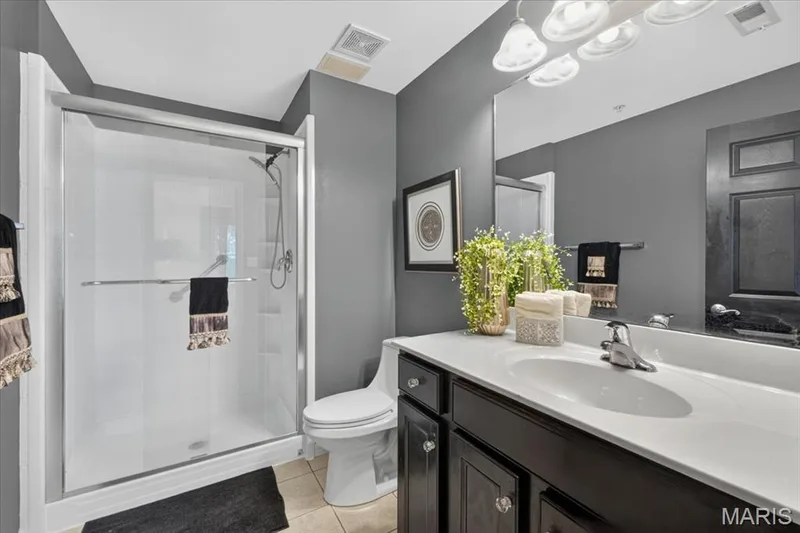 Bathroom featuring a glass-enclosed shower, a vanity with a white countertop and dark cabinetry, a large wall-mounted mirror, and tile flooring — 5 Monarch Trace Court #108, Chesterfield, MO 63017