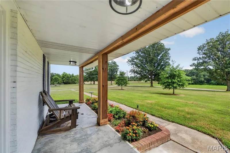 Covered porch featuring white brick exterior, exposed wood beam supports, concrete flooring, and an integrated flower bed with brick trim — 33 Timber Ridge Drive, Marthasville, MO 63357