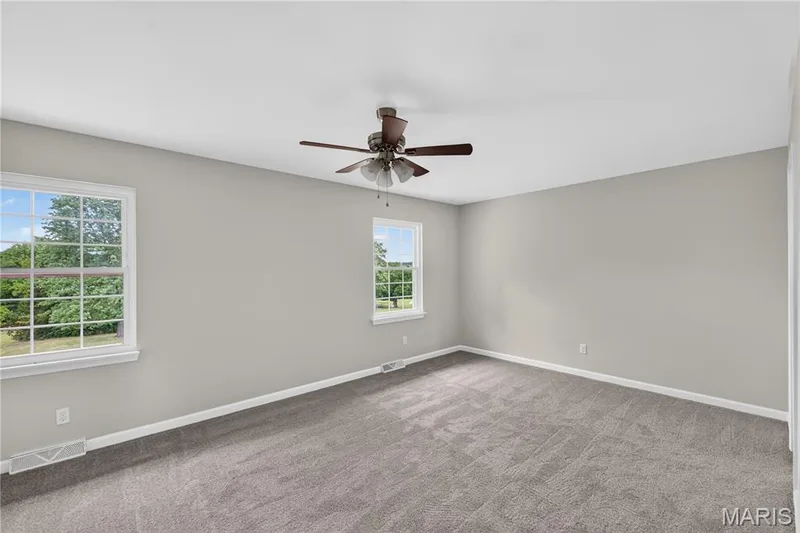 Spacious room featuring neutral grey wall paint, grey carpeting, two windows with white trim, and a ceiling fan with integrated lighting — 33 Timber Ridge Drive, Marthasville, MO 63357