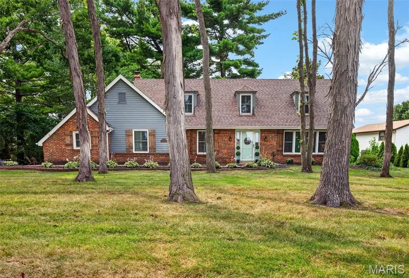 Brick and siding facade with dormer windows, featuring a light blue front door, decorative shutters, and mature trees — 4 Chantilly Court, Lake St. Louis, MO 63367