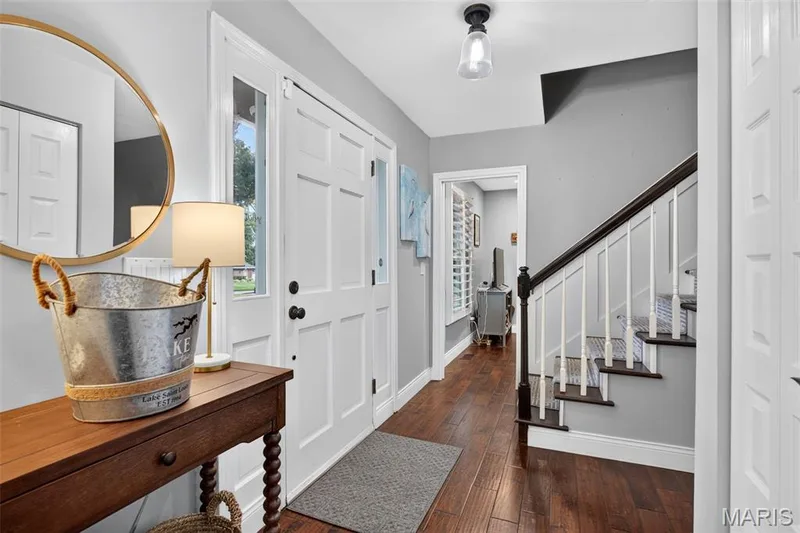 Entryway featuring wood-finish flooring, a paneled front door with sidelights, a contemporary flush mount light fixture, and a staircase with white risers and a dark wood handrail — 4 Chantilly Court, Lake St. Louis, MO 63367