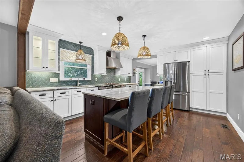 Spacious kitchen featuring an expansive center island with seating, white shaker cabinetry, stainless steel appliances, herringbone tile backsplash, and wood-finish flooring — 4 Chantilly Court, Lake St. Louis, MO 63367