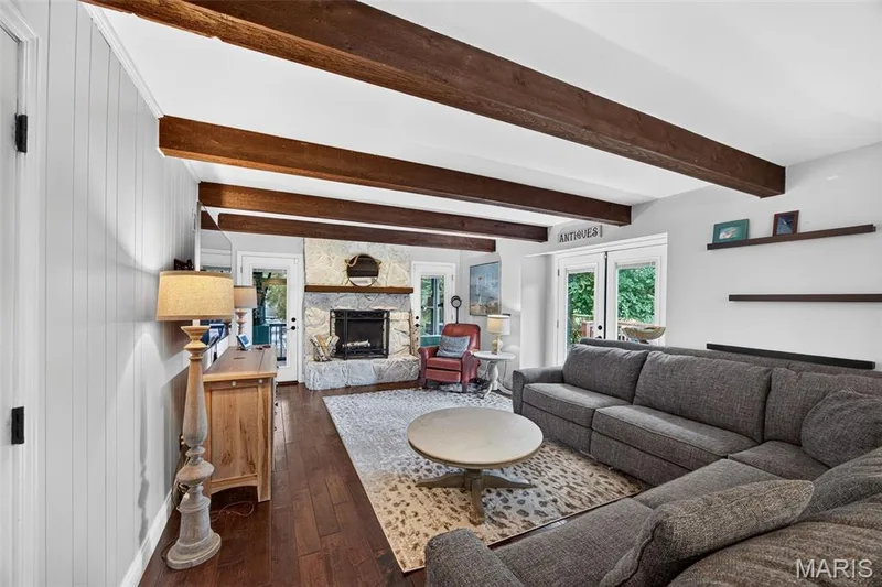 Living area featuring exposed wood ceiling beams, wood-finish flooring, a stone fireplace with hearth, and white shiplap paneling — 4 Chantilly Court, Lake St. Louis, MO 63367