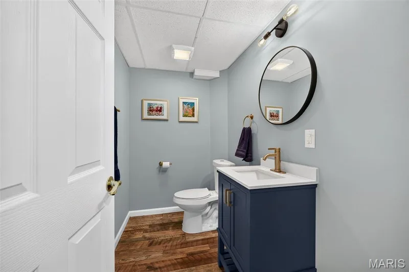 Bathroom featuring a blue vanity with a white countertop and brass-finish faucet, a round black-framed mirror, wood-finish flooring, and light blue walls — 4 Chantilly Court, Lake St. Louis, MO 63367