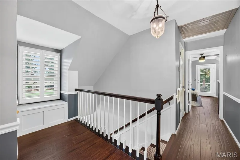 Upper-level hallway featuring wood-finish flooring, a built-in window bench with plantation shutters, a staircase with white spindles and a dark wood handrail, an overhead lantern-style light fixture, and a two-tone wall treatment — 4 Chantilly Court, Lake St. Louis, MO 63367