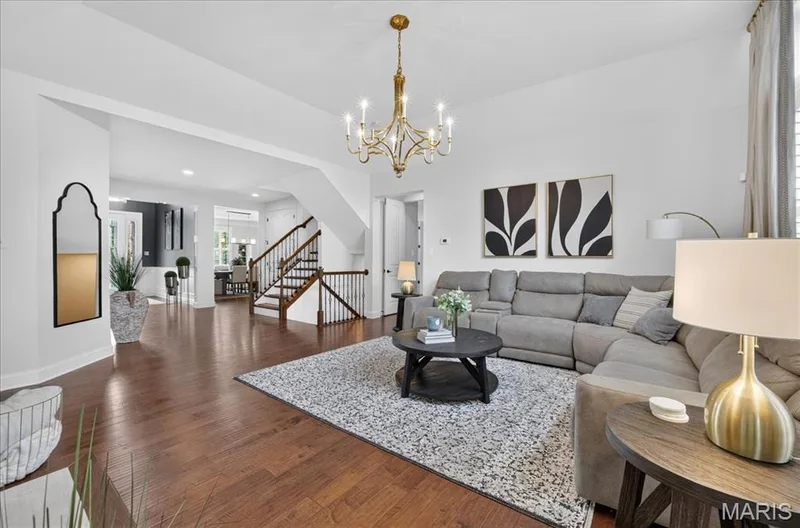 Spacious living area featuring rich wood-finish flooring, a traditional chandelier, and a prominent staircase with dark wood handrails and balusters — 302 Oak Stand Court, Chesterfield, MO 63005