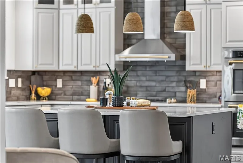 Modern kitchen featuring a dark gray stone-finish backsplash, light gray cabinetry with gold-tone hardware, and a center island with a light-toned countertop — 302 Oak Stand Court, Chesterfield, MO 63005
