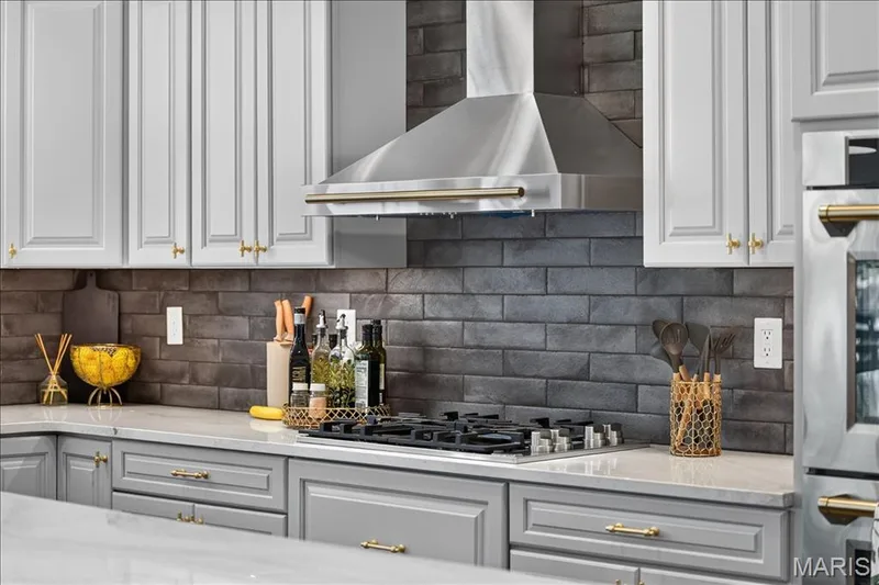 Gourmet kitchen featuring a stainless steel range hood, built-in gas cooktop, gray subway tile backsplash, white shaker-style upper cabinetry, and gray shaker-style lower cabinetry with gold-tone hardware — 302 Oak Stand Court, Chesterfield, MO 63005