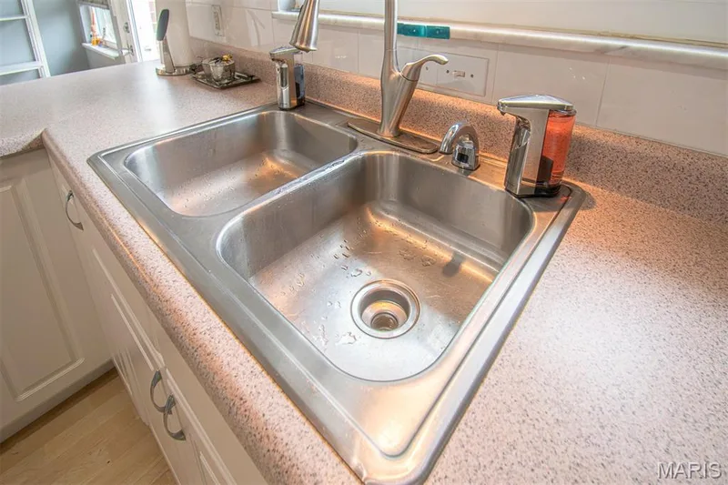 KITCHEN - Double basin stainless steel sink with a high-arc faucet, set into a speckled countertop with a white tiled backsplash, complemented by white cabinetry with dark hardware — 13261 COCHERO Drive, Chesterfield, MO 63141