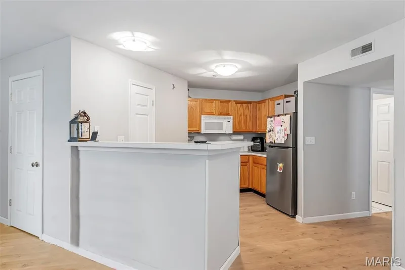 Open-concept kitchen featuring wood-finish flooring, light wood cabinetry, white countertops, and a stainless steel refrigerator — 640 Homerun Drive #37N, O'Fallon, MO 63366