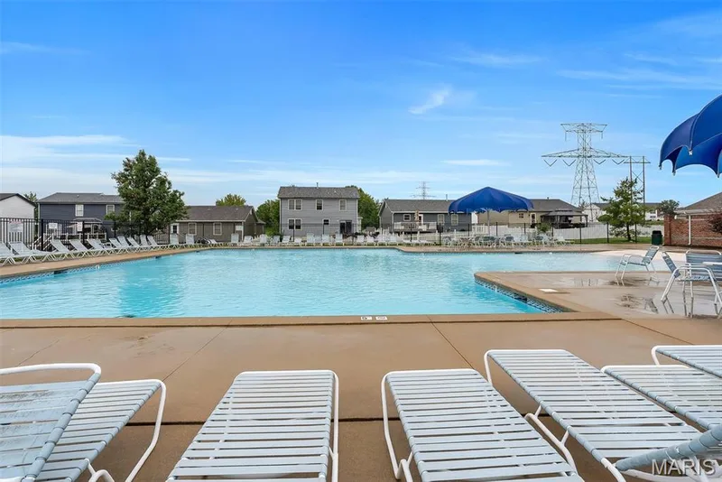 Expansive outdoor swimming pool featuring a large concrete deck, perimeter fencing, multiple lounge chairs, and blue shade umbrellas — 640 Homerun Drive #37N, O'Fallon, MO 63366