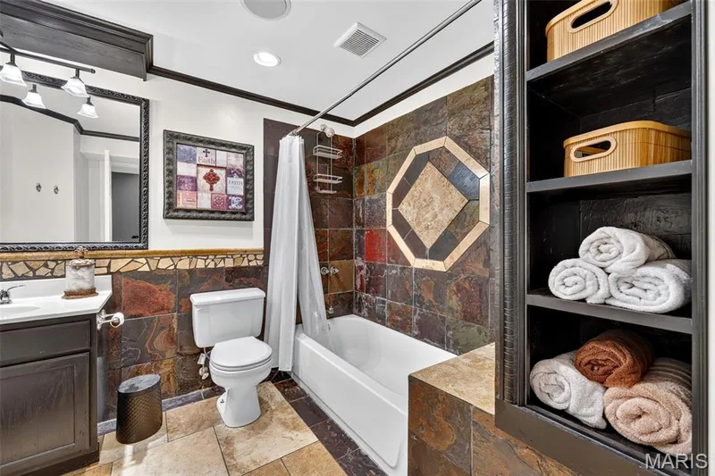 Built-in shelving, white vanity with integrated sink, rectangular mirror, full bathtub with shower, and multi-toned tile walls and flooring — 708 Woodland Ridge Drive, St Peters, MO 63376