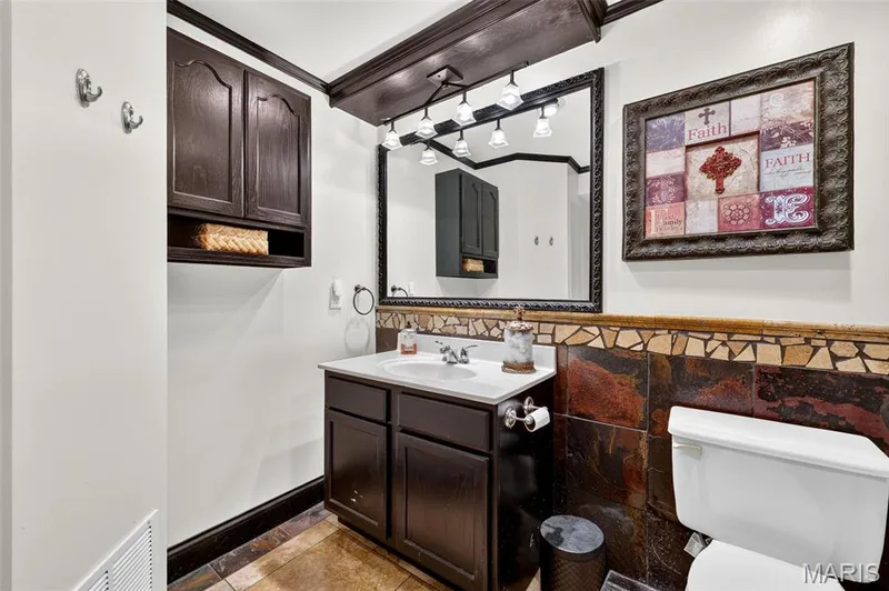 Dark wood-finish cabinetry, vanity with white basin, framed mirror, wall-mounted light fixture, and mosaic tile wainscoting — 708 Woodland Ridge Drive, St Peters, MO 63376