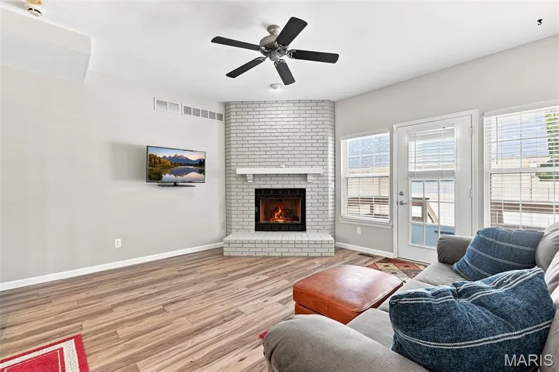 Wood-finish flooring throughout, ceiling fan with light fixture, corner brick fireplace with mantel, white door with glass panels, and multiple windows providing natural light — 16561 Carriage View Court, Wildwood, MO 63040