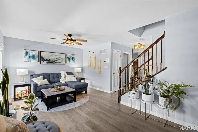 Open concept living area featuring wood-finish flooring, a ceiling fan with integrated lighting, and a prominent staircase with a wood handrail and balusters — 349 Shady Rock Lane, O'Fallon, MO 63368