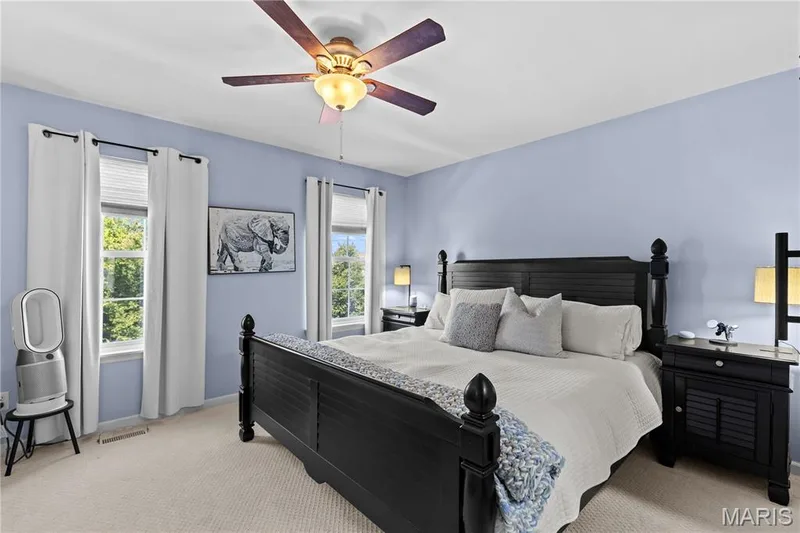 Bedroom featuring light blue walls, two windows with white blinds and curtains, a ceiling fan with light fixture, and plush carpeting — 349 Shady Rock Lane, O'Fallon, MO 63368