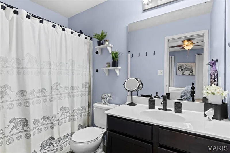 Full bathroom featuring a dark wood-finish vanity with a white countertop and integrated sink, matte black faucet, full-width wall mirror, and light blue wall paint — 349 Shady Rock Lane, O'Fallon, MO 63368