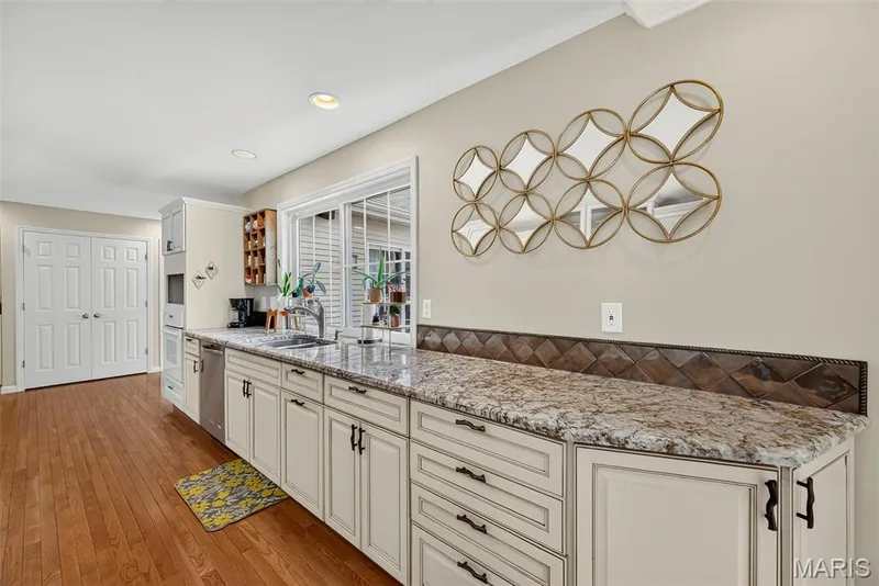 Kitchen featuring wood-finish flooring, light-toned cabinetry with dark hardware, granite-style countertops, a stainless steel sink, and a tiled backsplash — 15571 Rose Gate Lane, Chesterfield, MO 63017