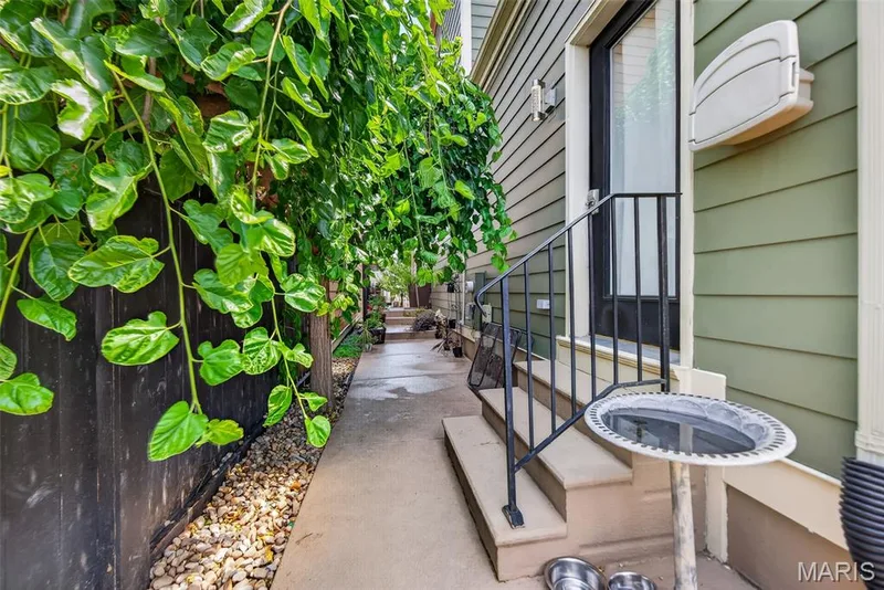 Charming side passage featuring lush green climbing ivy and mature foliage along a dark accent wall, concrete walkway with gravel base, black metal railings, and direct access to the residence — 3471 Hiram Street, St Charles, MO 63301
