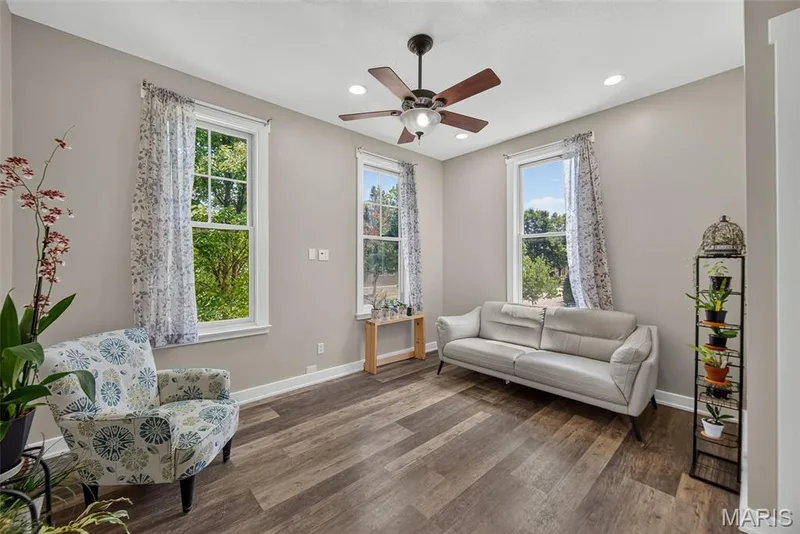 Bright and airy living room showcasing hardwood-finish flooring, multiple large windows with natural light, and a ceiling fan with integrated lighting — 3471 Hiram Street, St Charles, MO 63301
