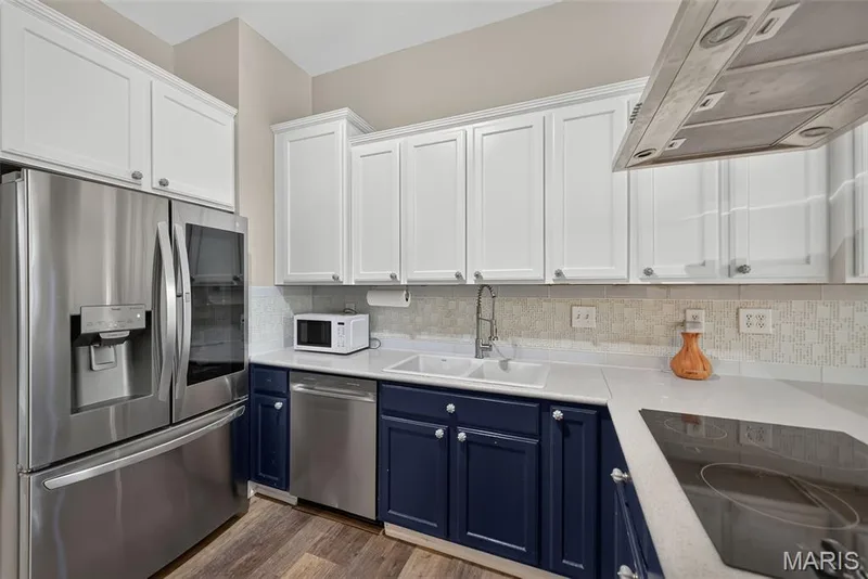 Modern kitchen featuring white shaker-style cabinetry paired with navy blue lower cabinets, stainless steel appliances including a French-door refrigerator and built-in microwave, quartz countertops, subway tile backsplash, and wood-finish flooring throughout — 3471 Hiram Street, St Charles, MO 63301