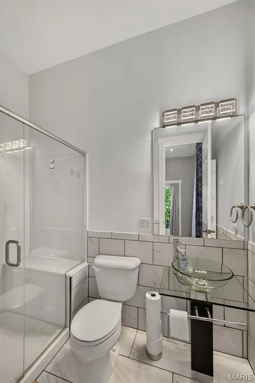 Modern bathroom featuring a clean-lined layout with subway tile wainscoting, a framed mirror with integrated lighting above the vanity, a ceramic vessel sink with contemporary faucet, and a separate glass-enclosed shower stall with frameless enclosure and tile surround — 3471 Hiram Street, St Charles, MO 63301