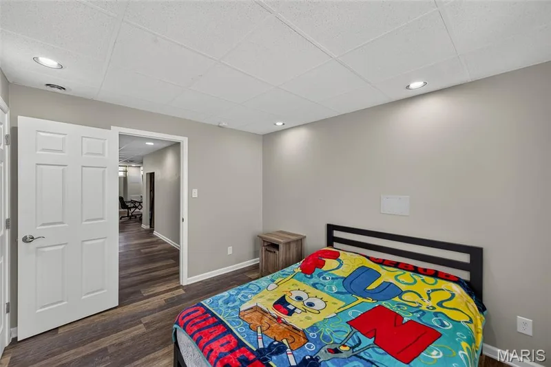 Finished basement bedroom featuring a queen-size bed frame with black metal headboard, wood-finish flooring throughout, recessed ceiling lighting, and direct access to an adjoining hallway — 3471 Hiram Street, St Charles, MO 63301