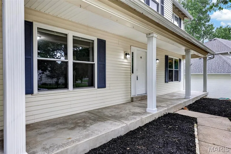 Covered front porch featuring white siding, dark blue shutters, white columns, and a concrete walkway — 2947 Bromley Drive, O'Fallon, MO 63368