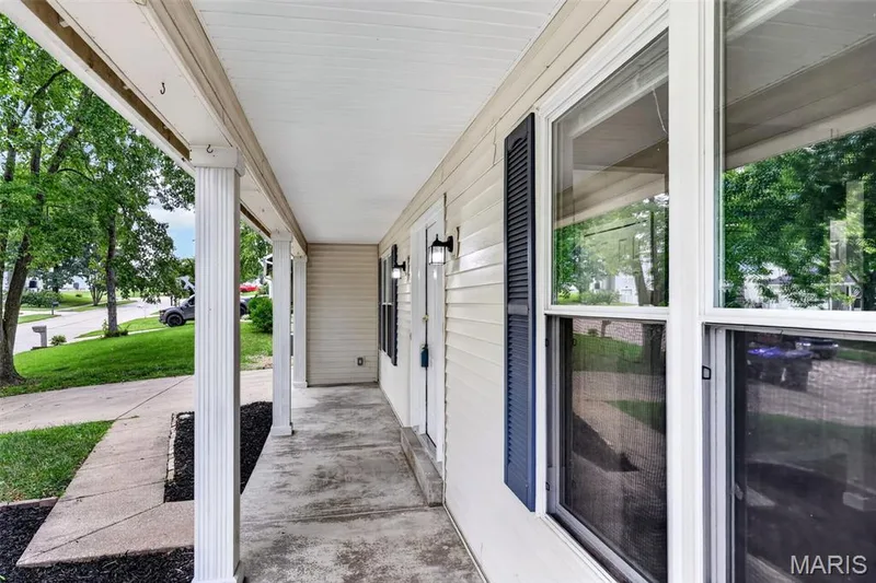 Welcoming covered entry featuring horizontal vinyl siding, large double-hung windows with screens, black hardware accents, and a concrete walkway lined with manicured landscaping and mature shade trees — 2947 Bromley Drive, O'Fallon, MO 63368