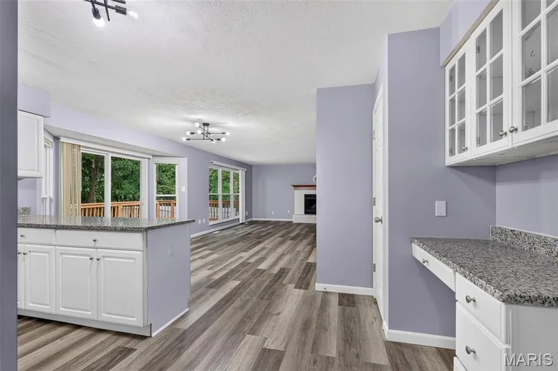 Open-concept living area featuring wood-finish flooring, a kitchen island with a granite-style countertop, white cabinetry with glass-front uppers, and multiple sliding glass doors leading to an exterior deck — 2947 Bromley Drive, O'Fallon, MO 63368