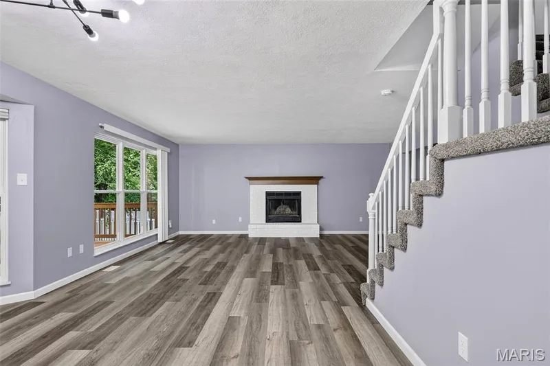 Spacious living area featuring wood-finish flooring, a fireplace with a white brick surround and wood mantel, and large windows with a deck beyond — 2947 Bromley Drive, O'Fallon, MO 63368