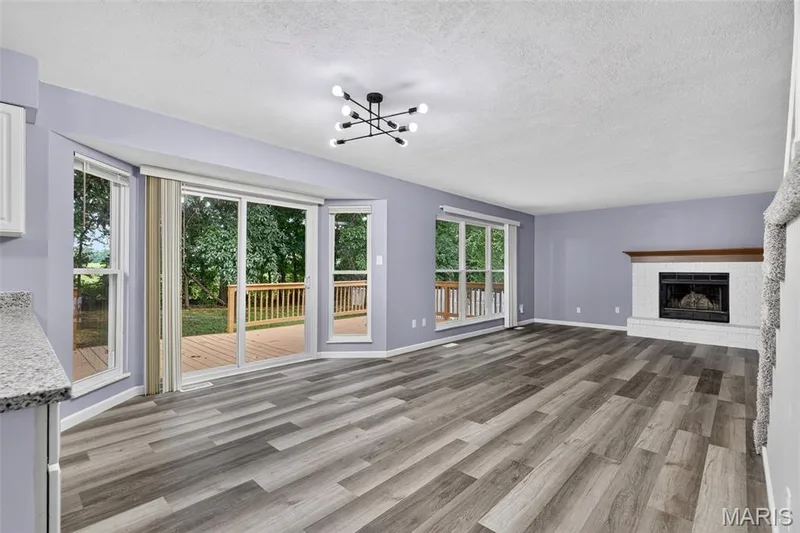 Spacious interior featuring wood-finish flooring, a contemporary chandelier, a white brick fireplace with a wood mantle, and sliding glass doors with surrounding windows leading to an exterior deck — 2947 Bromley Drive, O'Fallon, MO 63368