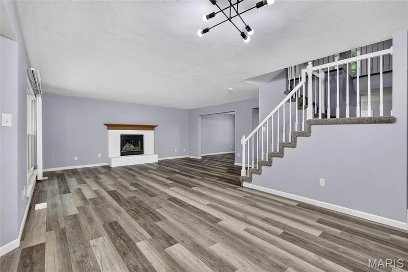 Spacious living area featuring wood-finish flooring, a white-painted brick fireplace with a wood mantle, and a contemporary chandelier — 2947 Bromley Drive, O'Fallon, MO 63368