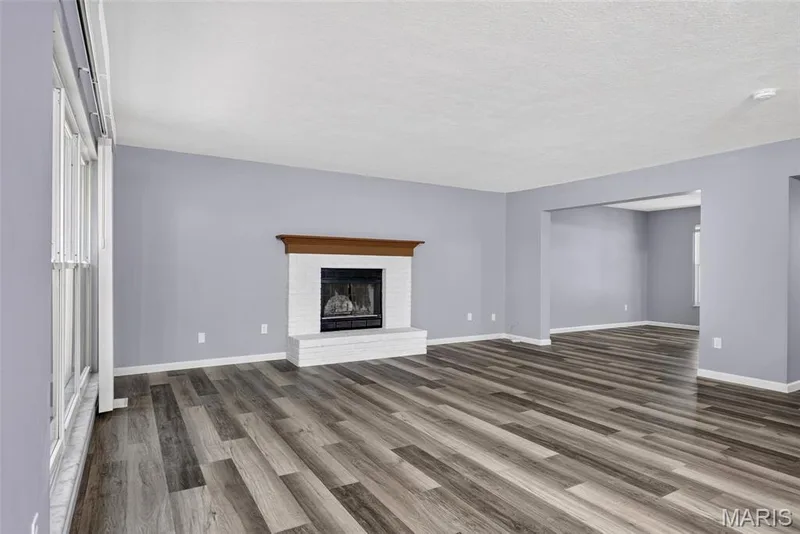 Spacious living area featuring multi-tone wood-finish flooring, a white brick fireplace with a wood mantel, and large windows — 2947 Bromley Drive, O'Fallon, MO 63368
