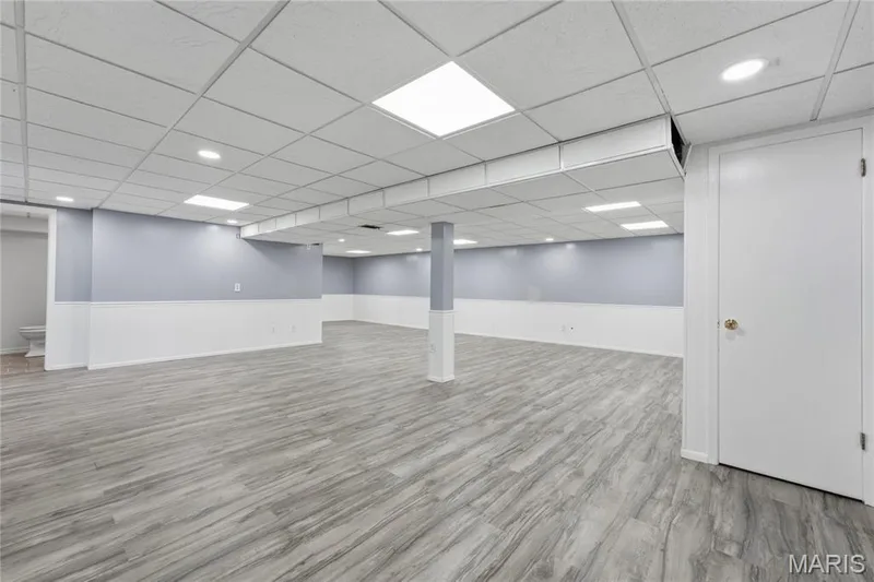 Expansive finished lower level featuring wood-finish flooring, a drop-ceiling with recessed lighting and square panel lights, two-tone walls with wainscoting, and a white interior door with a brass-finish knob — 1613 Tamarack Drive, St Charles, MO 63301