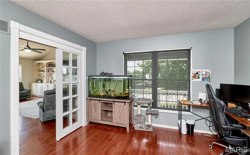 Room featuring wood-finish flooring, light blue wall paint, a window with exterior railing visible, and white French doors opening to an additional room with built-in shelving — 3 Floral Court, O'Fallon, MO 63368