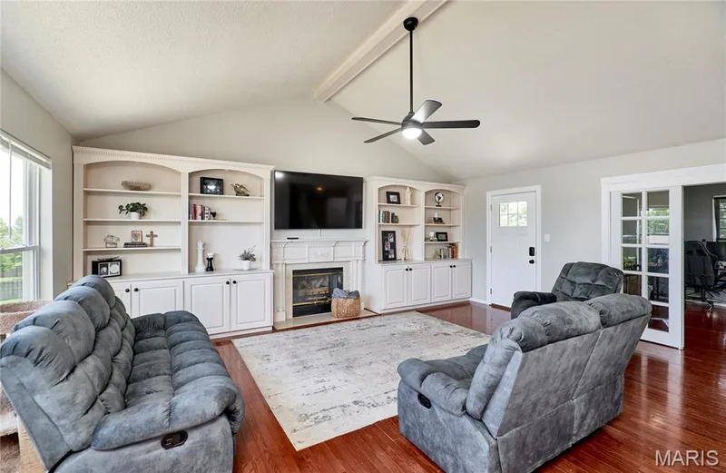 Living area featuring vaulted ceilings, a central fireplace with an integrated entertainment center, built-in shelving, rich wood-finish flooring, and a ceiling fan with integrated lighting — 3 Floral Court, O'Fallon, MO 63368