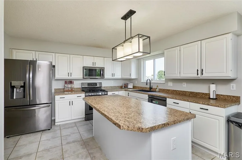 Kitchen featuring a central island with a speckled countertop, white cabinetry with dark hardware, stainless steel appliances, and a modern rectangular light fixture — 3 Floral Court, O'Fallon, MO 63368
