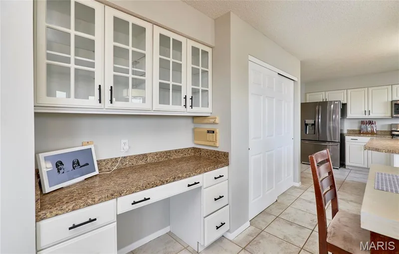 Bright kitchen workspace featuring white cabinetry with glass-front upper cabinets, a granite-style countertop, stainless steel appliances, and black hardware accents — 3 Floral Court, O'Fallon, MO 63368