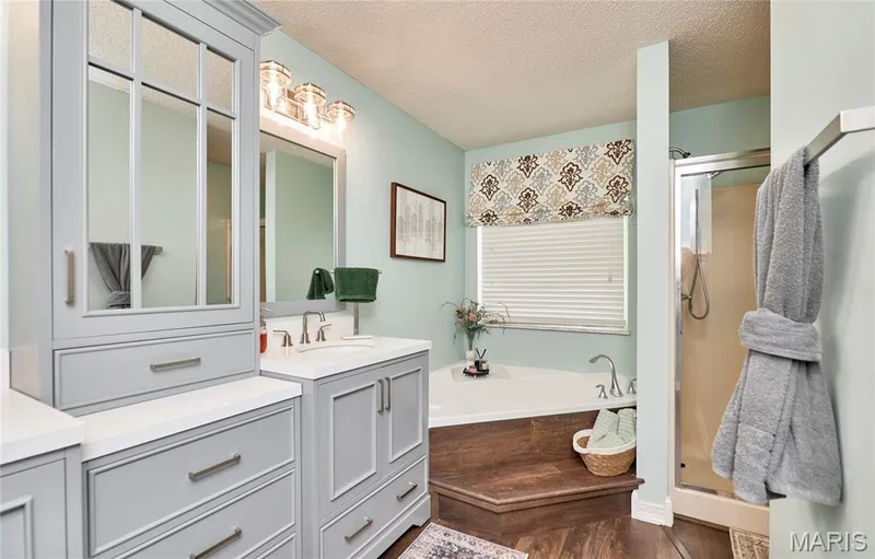 Bathroom featuring a corner soaking tub with wood-finish surround, glass-enclosed shower, and a light gray vanity with a white countertop — 3 Floral Court, O'Fallon, MO 63368
