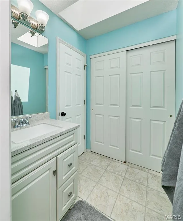 Bathroom featuring a vanity with an integrated sink and light-colored countertop, white cabinetry with silver-tone hardware, a wall-mounted mirror with an overhead light fixture, and light-toned tile flooring — 3 Floral Court, O'Fallon, MO 63368