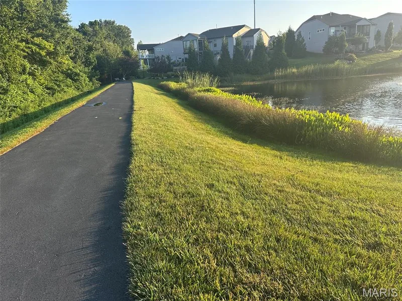 Paved pathway bordered by manicured lawn and mature trees, with a water feature and residential structures in the background — 110 Alta Mira Court, Dardenne Prairie, MO 63368