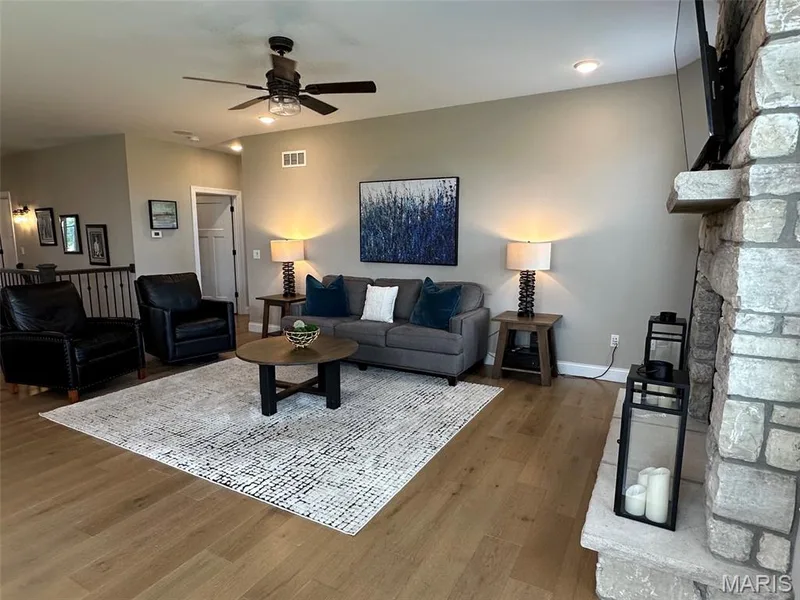 Spacious living area featuring light wood-finish flooring, a stone-clad fireplace with a stone mantel, a ceiling fan with integrated lighting, and recessed ceiling lights — 110 Alta Mira Court, Dardenne Prairie, MO 63368