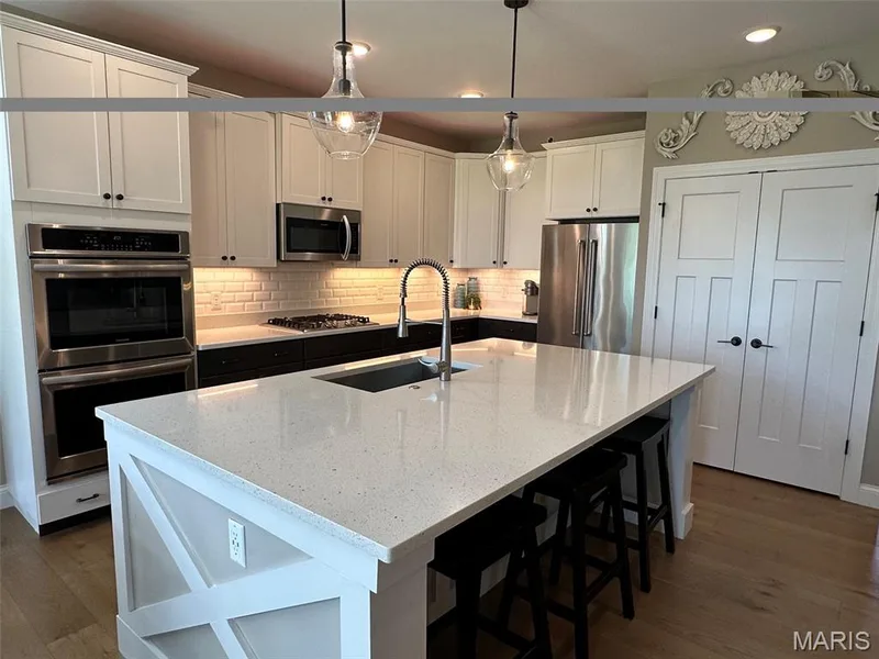 Kitchen featuring an expansive center island with a light-toned countertop, contrasting dark base cabinetry, a subway tile backsplash, stainless steel appliances, and wood-finish flooring — 110 Alta Mira Court, Dardenne Prairie, MO 63368